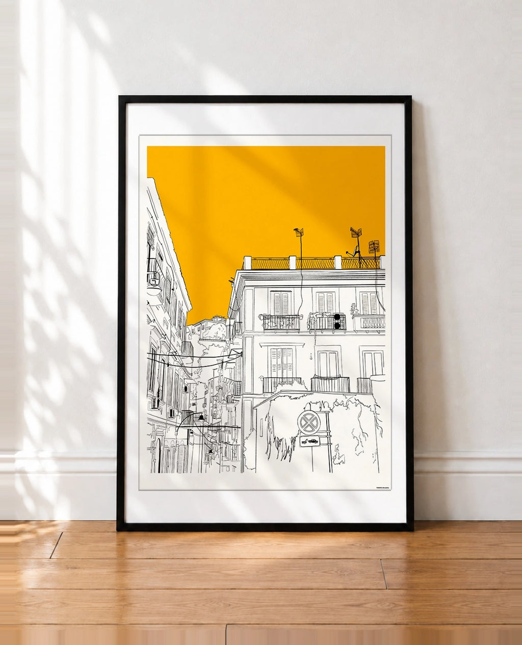A framed art print featuring a minimalist black line drawing of a street scene in Naples with a bold yellow sky, leaning against a white wall on a wooden floor.