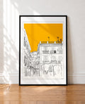 A framed art print featuring a minimalist black line drawing of a street scene in Naples with a bold yellow sky, leaning against a white wall on a wooden floor.