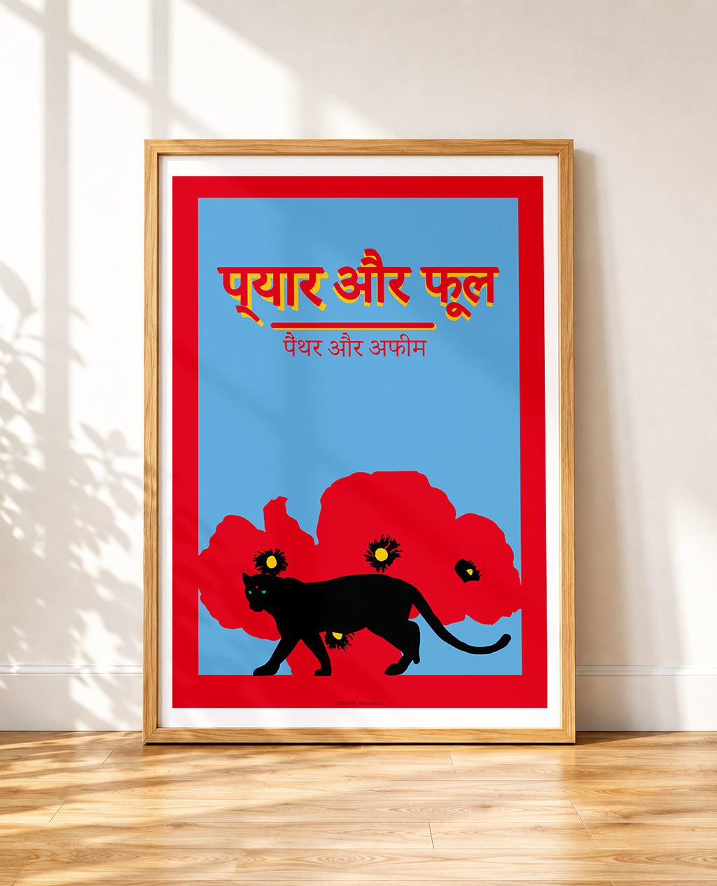 A framed poster leaning against a white wall on a wooden floor, featuring a blue background with a red border, Hindi text in orange and white, a black cat silhouette, and red flower shapes.