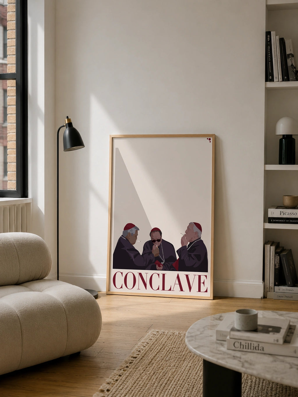 Framed Conclave poster in a light wood frame leaning against a beige wall in a bright living room, surrounded by a bouclé sofa, a black floor lamp and a bookshelf
