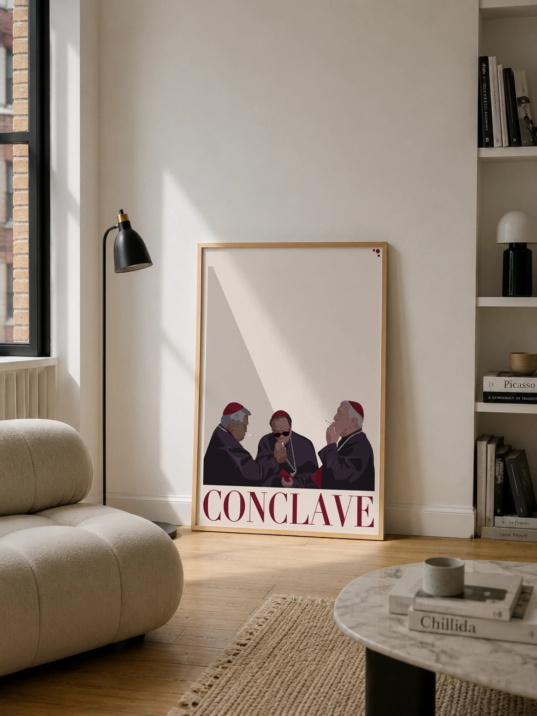 Framed Conclave poster in a light wood frame leaning against a beige wall in a bright living room, surrounded by a bouclé sofa, a black floor lamp and a bookshelf
