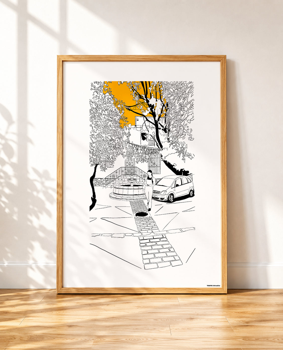framed Black and white line drawing of a cobblestone street scene with a stone fountain, a woman walking, a parked car and overhanging trees, with a bright orange sky breaking through the branches