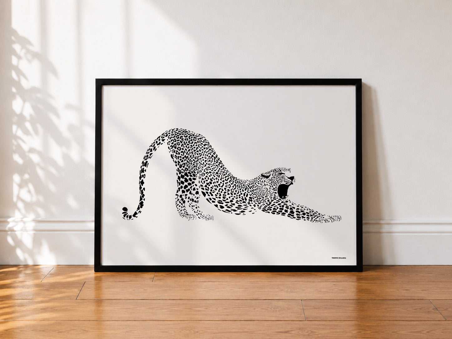 A framed black and white art print of a stretching leopard, leaning against a white wall on a wooden floor with soft leaf shadows.