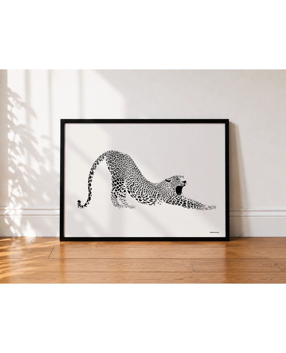 A framed black and white art print of a stretching leopard, leaning against a white wall on a wooden floor with soft leaf shadows.