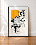 A framed art print featuring a black and white new york city street scene with a bold yellow block in the sky, leaning against a white wall on a wooden floor.