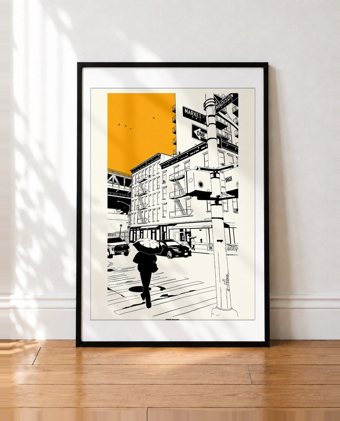 A framed art print featuring a black and white new york city street scene with a bold yellow block in the sky, leaning against a white wall on a wooden floor.