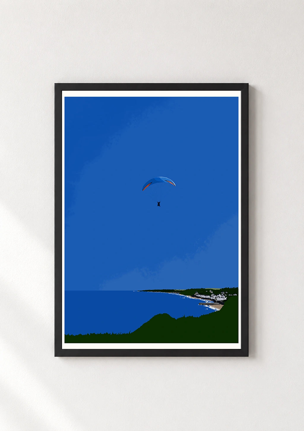Black framed print of a paraglider flying over a blue sky above a coastal cliff and village
