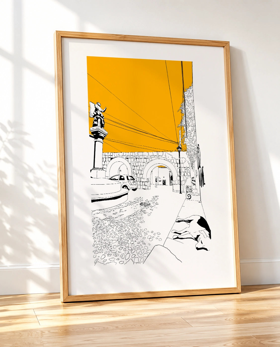 Framed black and white line-art poster leaning against a wall on a wooden floor, with natural light and tree shadows falling across the frame
