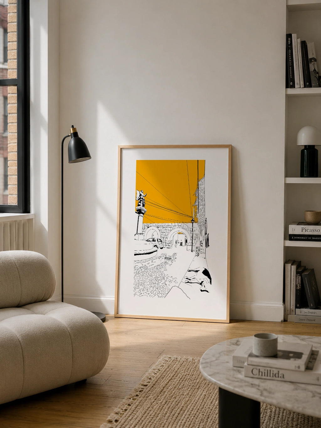 Framed black and white line-art poster leaning against a beige wall in a bright living room, surrounded by a bouclé sofa, a black floor lamp and a bookshelf
