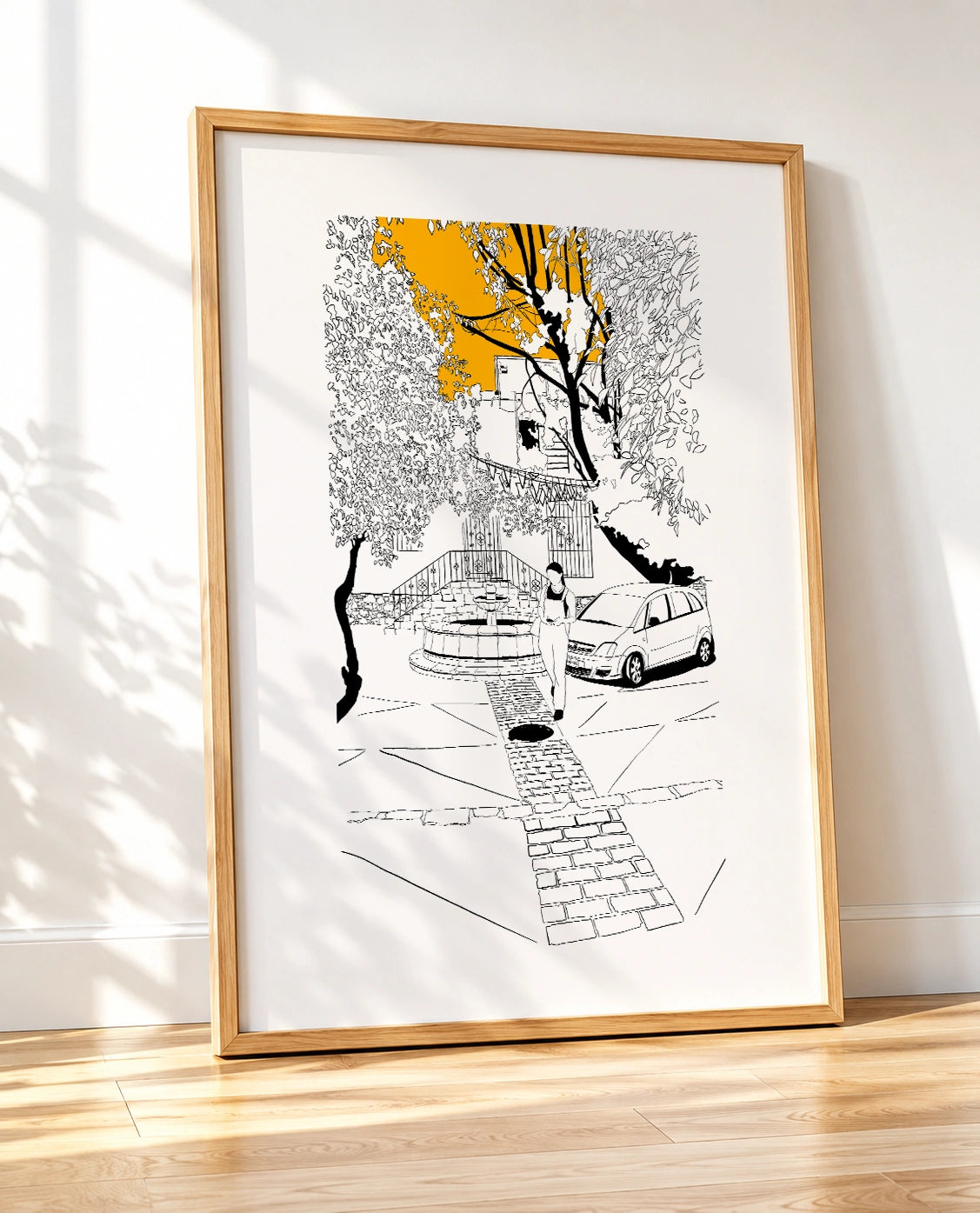 Framed black and white line-art poster leaning against a wall on a wooden floor, with natural light and tree shadows falling across the frame
