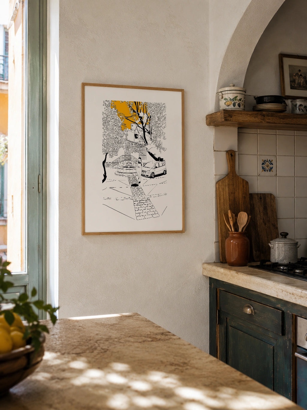 Framed black and white line-art poster hanging on a textured wall in a rustic Mediterranean kitchen with terracotta tiles, wooden utensils and a stone countertop
