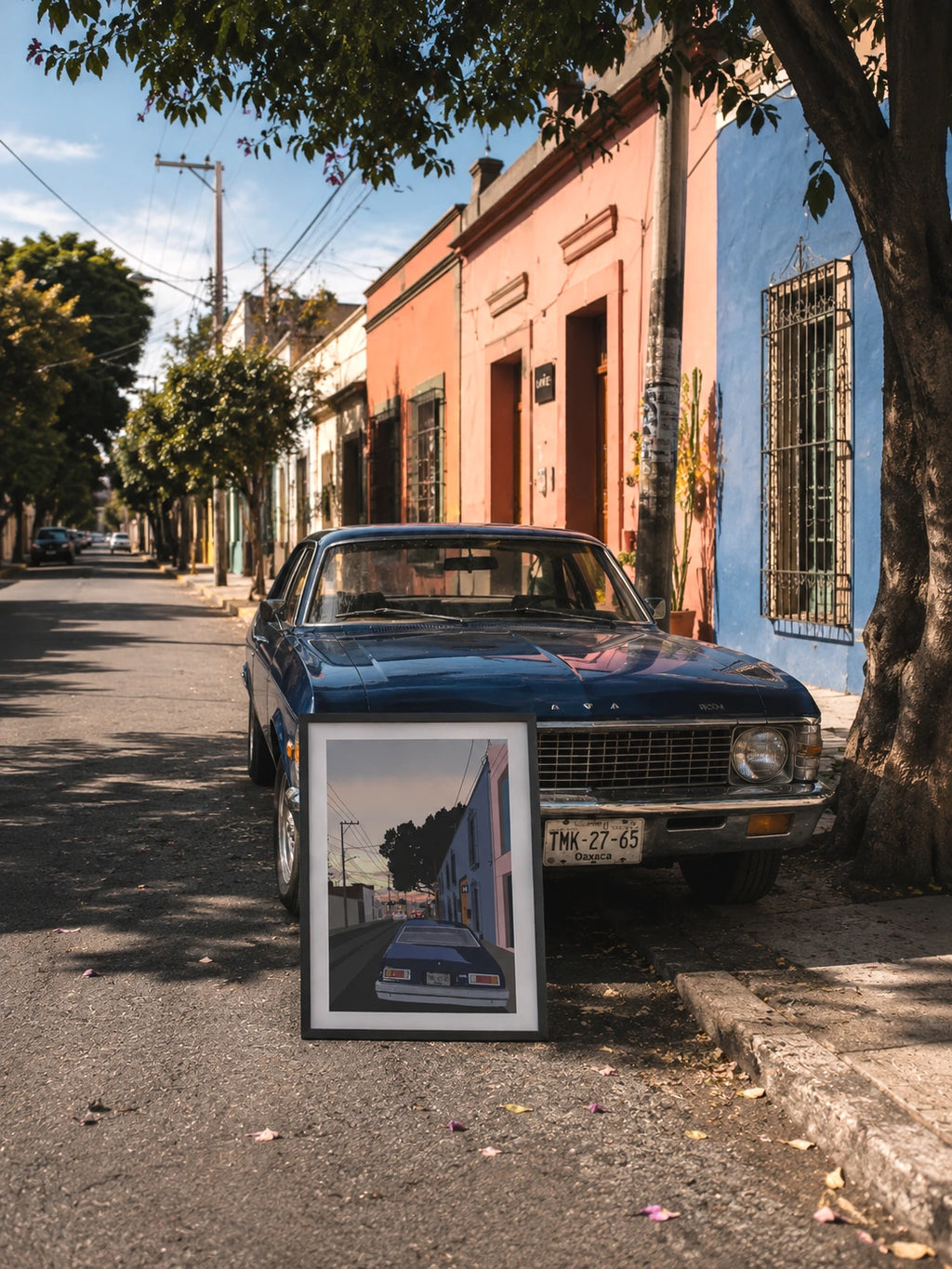 Framed Oaxaca Nova poster leaning against a blue Chevrolet Nova parked on a colorful cobblestone street in Oaxaca, in front of pink and blue facades under a tree
