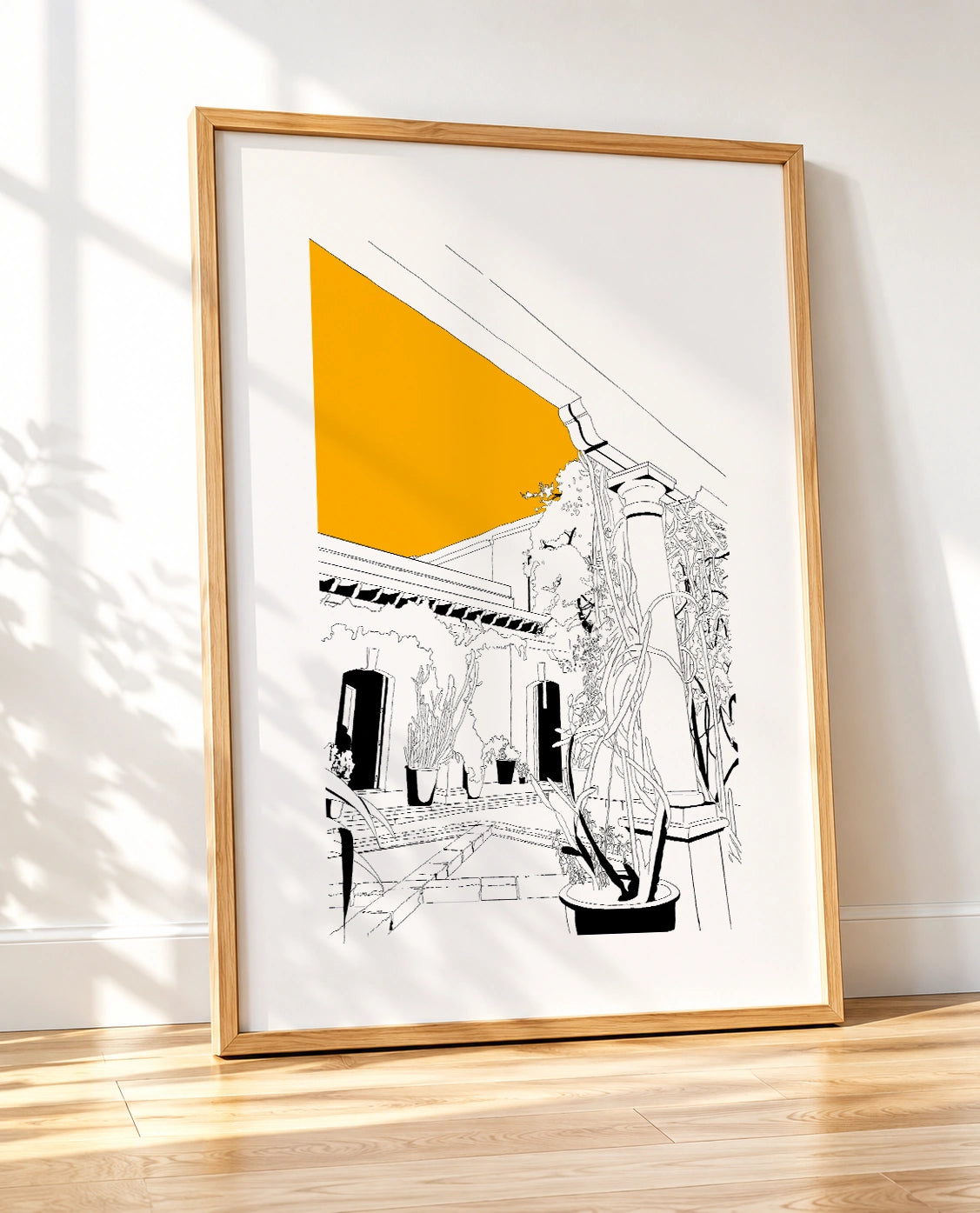 Framed black and white line-art poster leaning against a wall on a wooden floor, with natural light and tree shadows falling across the frame
