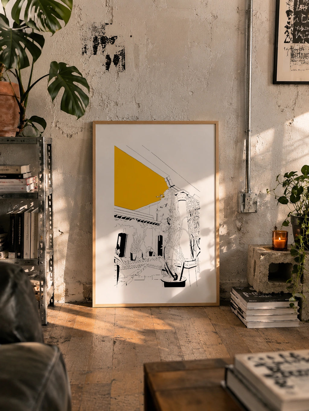 Framed black and white line-art poster leaning against a raw concrete wall in an industrial loft, surrounded by monstera plants, stacked magazines and a lit candle
