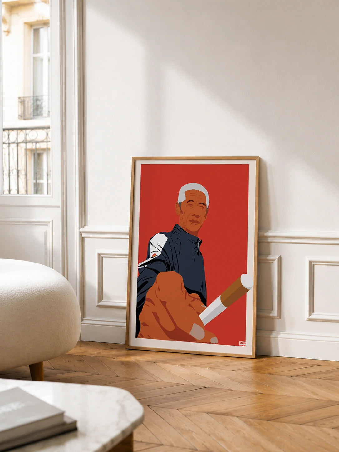 Framed Old Man Cigarette poster leaning against a wall in a Parisian-style room with chevron parquet flooring, wainscoting and a boucle ottoman, near tall windows with a wrought-iron balcony
