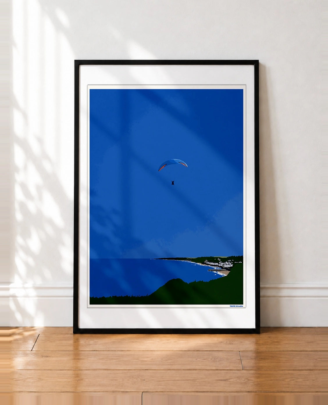 framed Illustration of a paraglider with a blue and orange canopy flying high above a coastline with cliffs and a village
