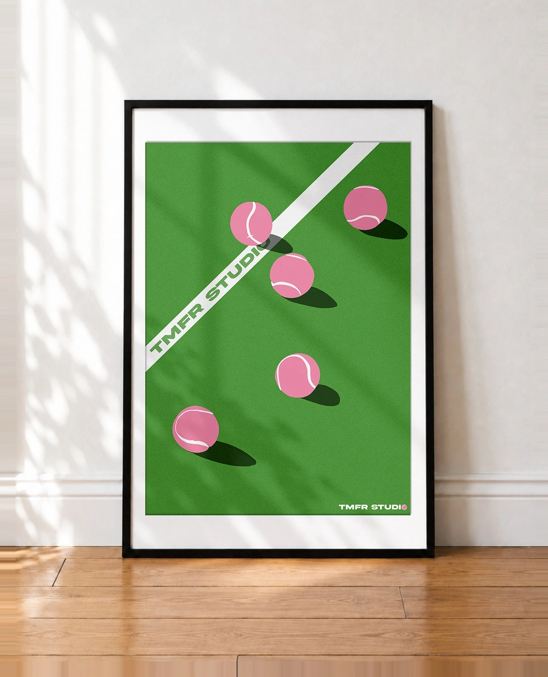A framed minimalist tennis-themed art print leaning against a white wall on a wooden floor. The print features a green court background with pink tennis balls, a white diagonal boundary line, and the text 'TMFR STUDIO' in green and white, enclosed in a black frame.