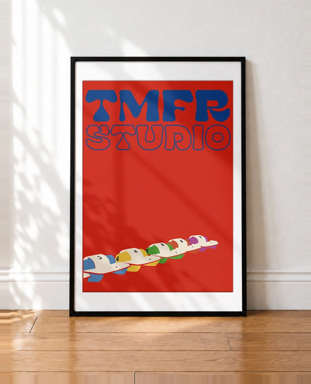 A framed art print leaning against a white wall on a wooden floor. The print features a bold red background with the words 'TMFR STUDIO' in blue retro lettering at the top and a row of colorful cartoon-style characters in a boat at the bottom.