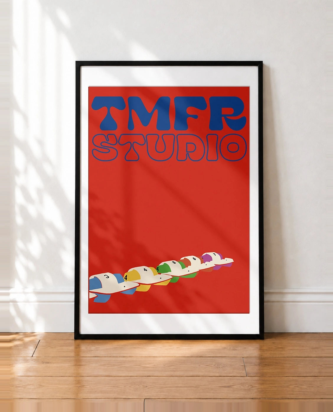 A framed art print leaning against a white wall on a wooden floor. The print features a bold red background with the words 'TMFR STUDIO' in blue retro lettering at the top and a row of colorful cartoon-style characters in a boat at the bottom.