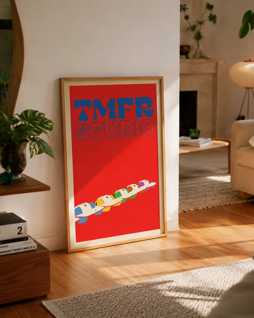 Framed TMFR Studio poster leaning against a wall near a fireplace, with a wavy wooden mirror, a monstera plant and stacked Magnum photography books nearby
